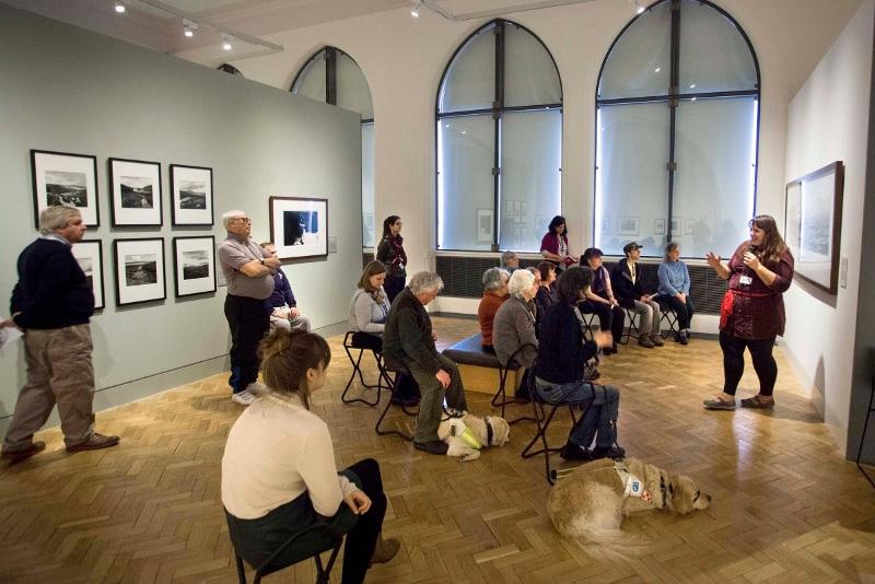 A group of visually impaired visitors listening to a talk given in the photography gallery. Two guide dogs are lying obediently on the floor.