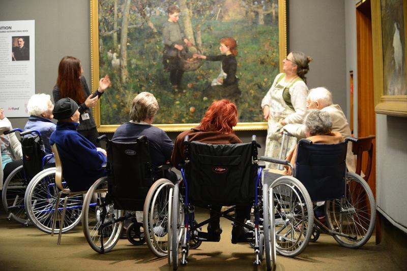 A woman in an apron standing in from of a painting by James Guthrie. The painting depicts a red-headed girl and a boy, the girl in placing an apple in the boy's basket. Gathered around the painting are a small group of wheelchair users listening to another woman describing the painting